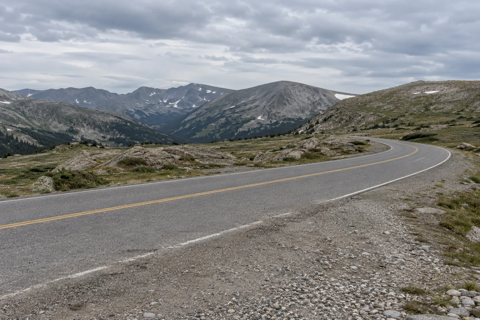 A paved road bends across treeless rocky tundra below grey mountain ridges and an overcast sky