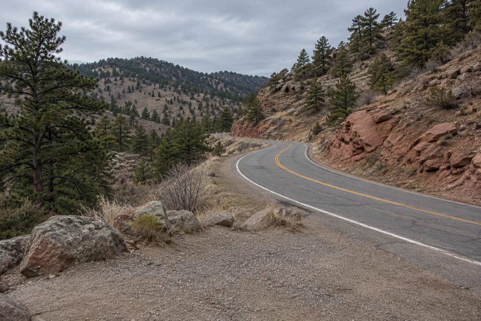 A paved road curves around rocky foothills and scattered pines beneath an overcast sky