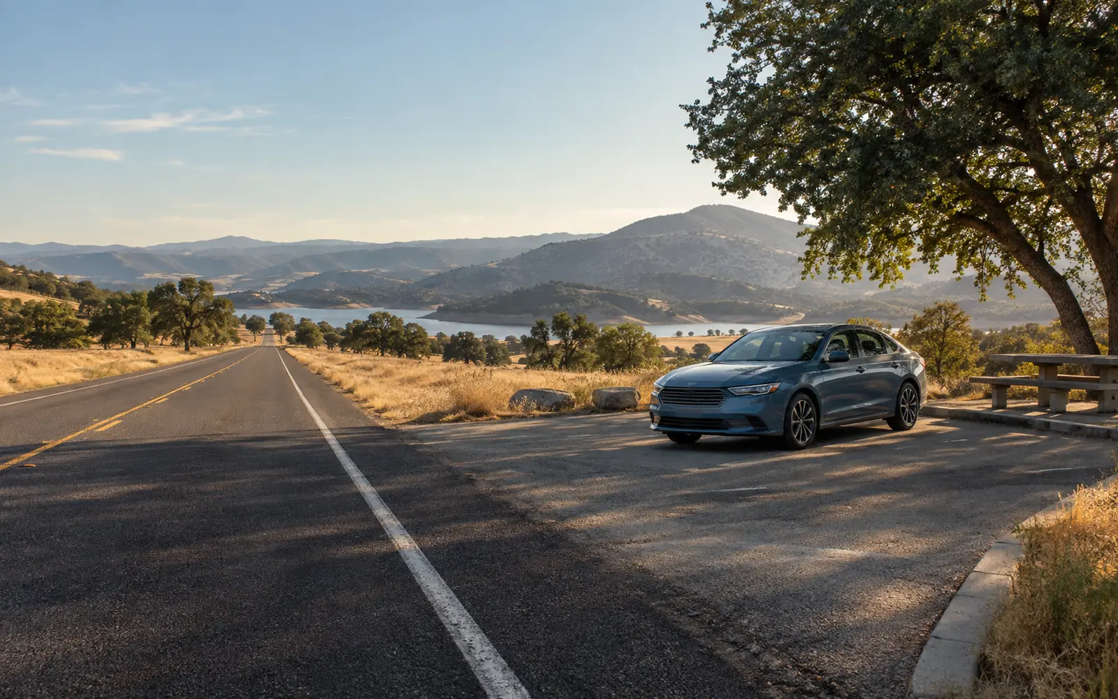 A two-lane highway stretching through open country in late afternoon light with an unbadged sedan parked at a rest area